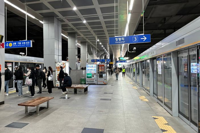 A Seoul subway platform with people seated and standing on a bench, platform screen doors, a blue directional sign reading Cheongnyangni, vending machines, and a train stopped alongside.