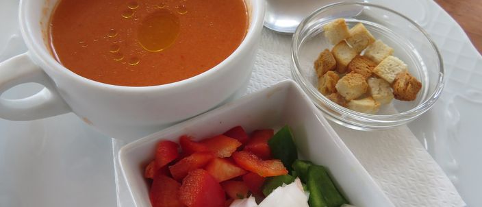 A white bowl of smooth, bright-red Andalusian gazpacho with a small dish of croutons and diced vegetables to stir in.