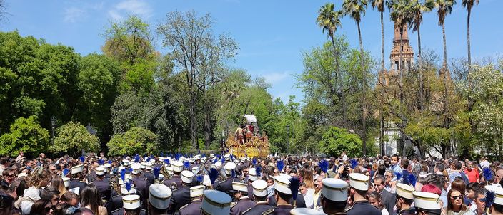 A Semana Santa procession in Parque de María Luisa: a hooded brass band in dark uniforms marching in the foreground, a gilded paso carried under palm trees, dense crowds lining the route in bright spring sunshine with a tiled tower behind.