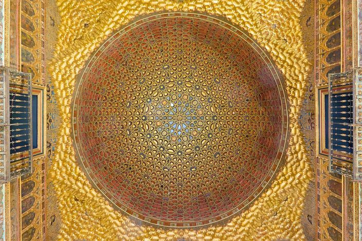 Looking straight up into a gilded wooden dome of interlocking star patterns in red, green and gold, framed by richly patterned gold walls with two shuttered windows below it on either side.