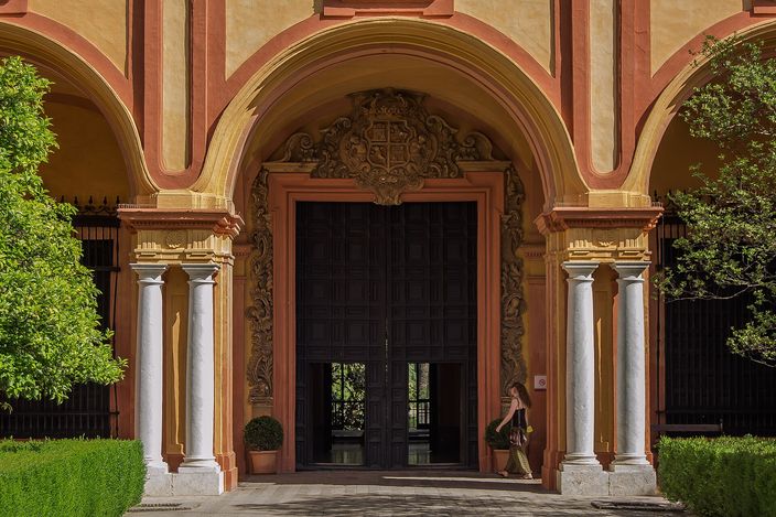 A gallery in the Alcazar of Seville: white marble columns and wide pink-and-ochre arches framing a carved stone doorcase with a coat of arms above tall dark timber doors, clipped hedges and greenery on either side and a single figure walking past.