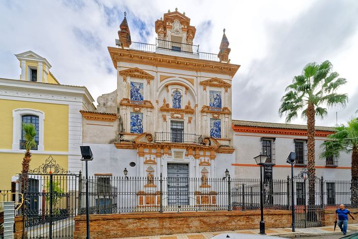 The white and ochre Baroque facade of the church of the Hospital de la Santa Caridad in Seville, with blue-and-white tiled panels of standing figures set between its pilasters, a wrought-iron railing and lamp posts in front of it and a palm tree rising beside it.