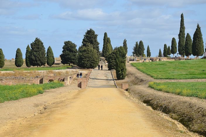 A broad flat Roman street at Italica paved in worn stone slabs, running away from the camera between lines of low brick pier stumps and dark cypress trees, with a few small figures walking in the distance under a blue sky.