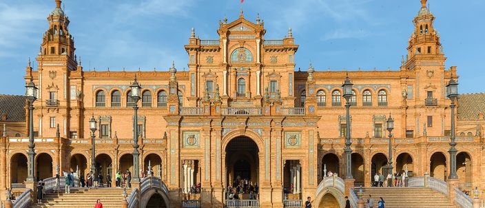 The main pavilion of Seville's Plaza de España in low golden sunlight: a long curved facade of orange brick and pale stone with a tall central gable, arcaded galleries on either side, square towers at each end, and ornamental balustraded bridges over the canal with people crossing them.
