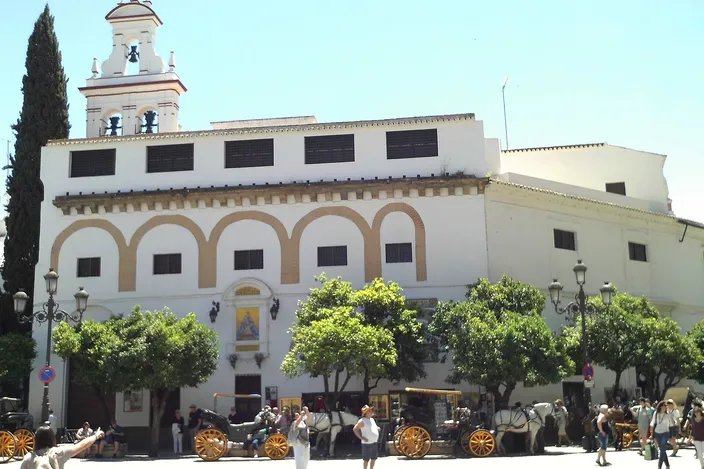 The whitewashed Convento de la Encarnación in Seville, with a tiled bell tower, arcaded upper facade and shady orange trees in front, horse-drawn carriages and pedestrians on the sunlit square below.