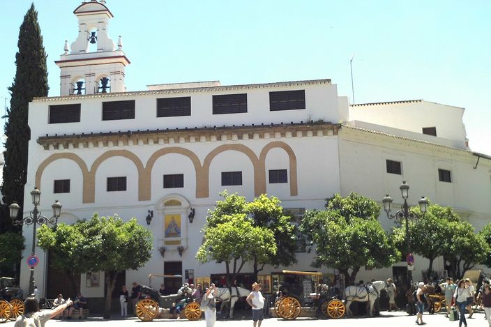 The whitewashed Convento de la Encarnación in Seville, with a tiled bell tower, arcaded upper facade and shady orange trees in front, horse-drawn carriages and pedestrians on the sunlit square below.