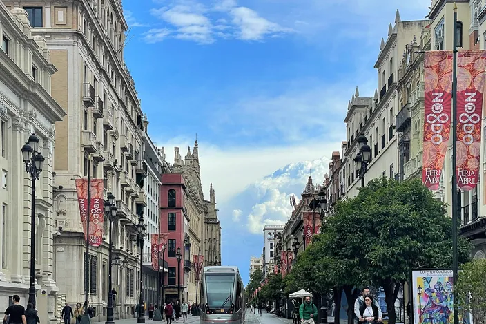 A modern MetroCentro tram running down a wide pedestrianized avenue in central Seville, grand pale stone buildings hung with Feria banners on either side and the cathedral's tower visible down the street under a bright blue sky