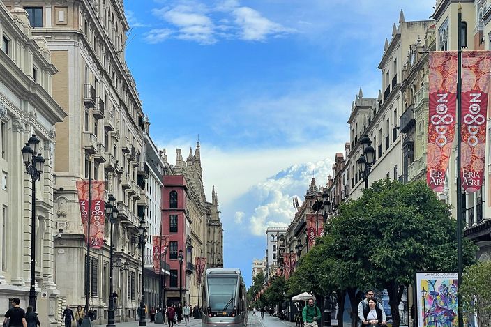 A modern MetroCentro tram running down a wide pedestrianised avenue in central Seville, grand pale stone buildings hung with Feria banners on either side and the cathedral's tower visible down the street under a bright blue sky