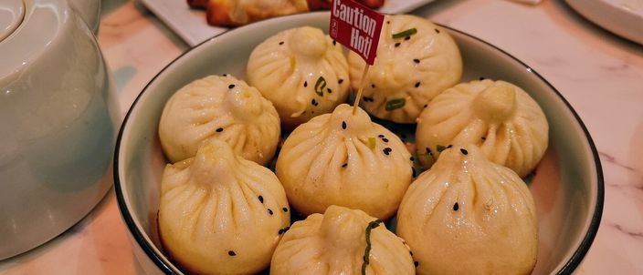 A bowl of plump pan-fried pork buns with pleated tops, scattered with sesame seeds and chopped spring onion.