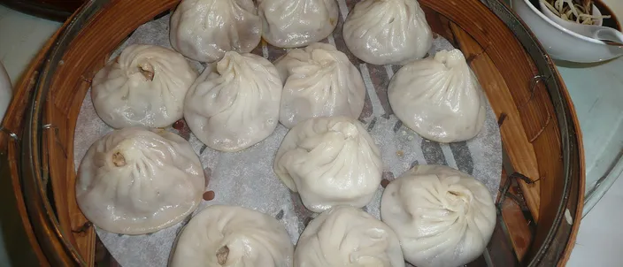 A round bamboo steamer filled with pale, translucent pleated xiaolongbao, each dumpling gathered into a topknot of dough.