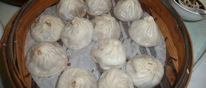 A round bamboo steamer filled with pale, translucent pleated xiaolongbao, each dumpling gathered into a topknot of dough.