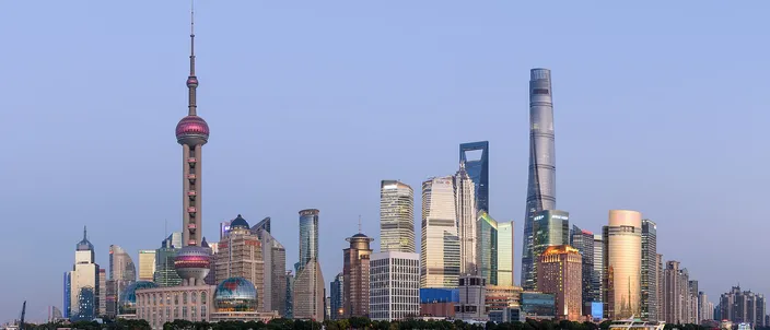 The Pudong skyline at dusk seen across the Huangpu River from the Bund — the Oriental Pearl Tower on the left, the twisting Shanghai Tower and the bottle-opener World Financial Center on the right, under a clear pale-blue sky.
