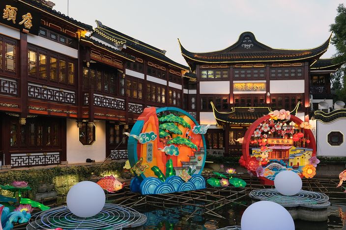 The Yu Garden bazaar at dusk, two storeys of dark timber with gold-trimmed upturned eaves lit from within, standing behind a pond in which large circular lantern sculptures of flowers, clouds and a landscape are moored.