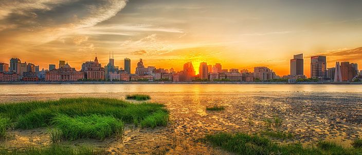 Sunset across the Huangpu River seen from a grassy tidal mudflat, with the low granite skyline of the Bund — a domed bank, a square clock tower and a green pyramid-roofed hotel among them — silhouetted against an orange sky on the far bank.