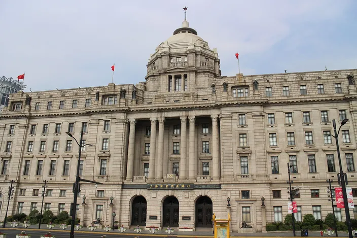 The stone facade of the former HSBC Building on the Bund in sunshine: a shallow dome on a drum above a portico of paired Corinthian columns, ranks of windows to either side, red flags along the parapet and three arched doorways at street level.