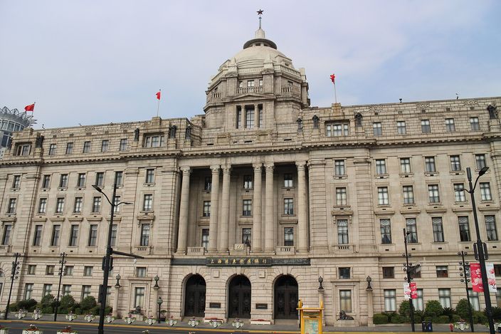 The stone facade of the former HSBC Building on the Bund in sunshine: a shallow dome on a drum above a portico of paired Corinthian columns, ranks of windows to either side, red flags along the parapet and three arched doorways at street level.