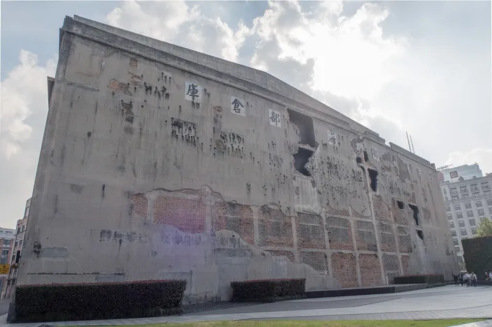 The west wall of the Sihang Warehouse, a gray concrete slab pitted all over with shell and bullet damage and torn by several ragged holes, standing above an empty paved square under a bright cloudy sky.
