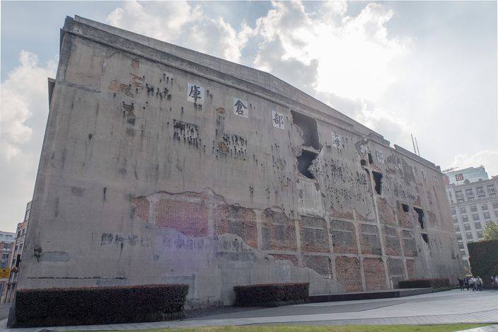 The west wall of the Sihang Warehouse, a grey concrete slab pitted all over with shell and bullet damage and torn by several ragged holes, standing above an empty paved square under a bright cloudy sky.