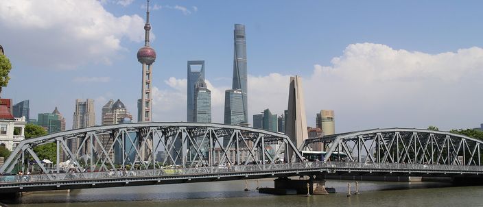 The grey steel truss of the Waibaidu Bridge spanning the mouth of Suzhou Creek in bright sunshine, with the Pudong towers behind it under a blue sky — the Oriental Pearl Tower's pink spheres on the left, the twisting Shanghai Tower and the bottle-opener World Financial Center to their right.
