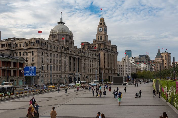 The Bund promenade in daytime, with the grey-domed former HSBC Building and the Customs House clock tower rising behind, people walking the wide riverside walkway