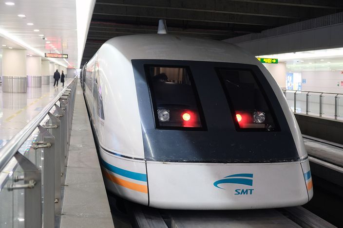 The white-and-blue Shanghai Maglev train stopped at a station platform, its aerodynamic nose facing the camera with red tail lights lit and the SMT logo below the windscreen