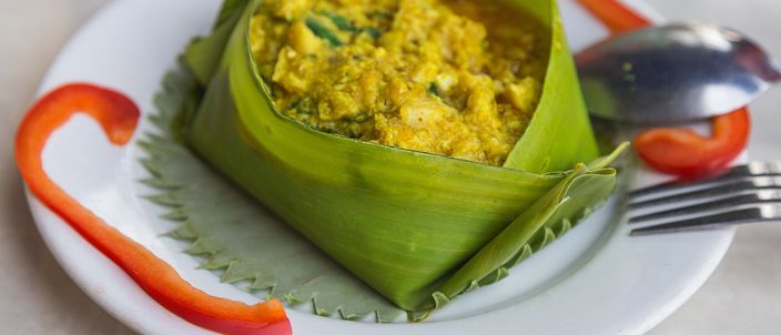 A serving of fish amok, a set yellow coconut-and-spice curry, steamed and presented in a folded banana-leaf cup on a white plate with curls of red chilli.
