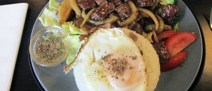 A plate of beef lok lak: seared cubes of beef with onions piled on a bed of lettuce, cucumber and tomato, topped with a fried egg dusted with cracked black pepper, with a small dish of lime-and-pepper dip alongside.