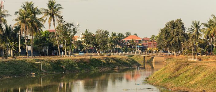 The Siem Reap River in daylight, calm brown water reflecting palm trees and grassy banks, with a low road bridge and low-rise town buildings under a warm hazy sky