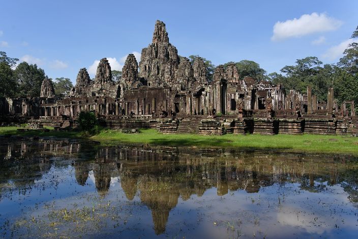 The face-towers of the Bayon rising in a dense grey stack above the temple's stepped terraces, framed by trees, with a green bank and a still pond in the foreground carrying the whole ruin in reflection under a blue sky with white cloud