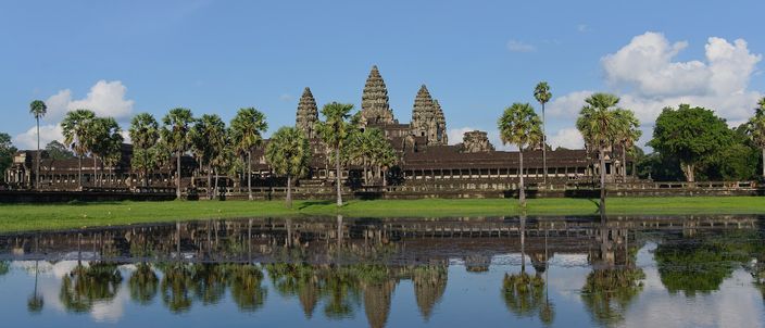 The five towers of Angkor Wat ranged along the horizon above the long low outer gallery, a line of palms and sugar palms in front of them, and the whole silhouette doubled in the still water of the reflecting pond that fills the lower half of the frame, under a blue sky with white cumulus