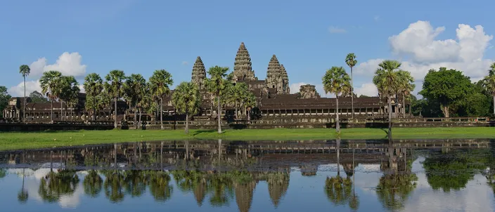 The five towers of Angkor Wat ranged along the horizon above the long low outer gallery, a line of palms and sugar palms in front of them, and the whole silhouette doubled in the still water of the reflecting pond that fills the lower half of the frame, under a blue sky with white cumulus