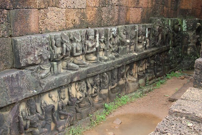 Tiers of carved figures -- seated kings, attendants and court women -- covering a stone wall that runs away from the camera at the Terrace of the Leper King, with a narrow paved gap and standing rainwater at its foot and grass growing from the joints