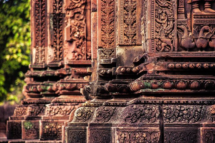 A close view of the rose-pink sandstone of Banteay Srei, two tower corners carved in dense foliate scrollwork with small figures and a bird set deep into the stone, the background dissolving into green