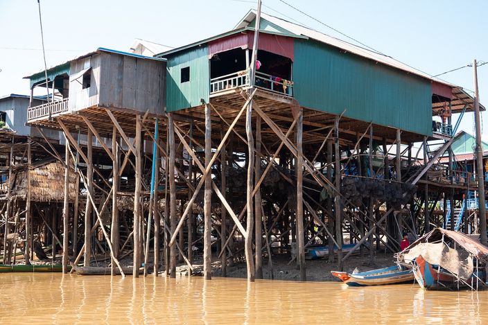 Wooden houses at Kampong Phluk standing on stilts three or four metres above brown floodplain water, their painted timber and corrugated walls raised on a forest of poles, with a narrow wooden boat moored at the right