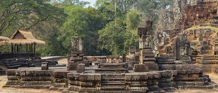 A broad terrace of stepped stone blocks inside the Baphuon enclosure at Angkor Thom, dry and empty in strong sun, with a thatched shelter on the left, the temple's weathered eastern stonework rising on the right and dense green forest closing the background
