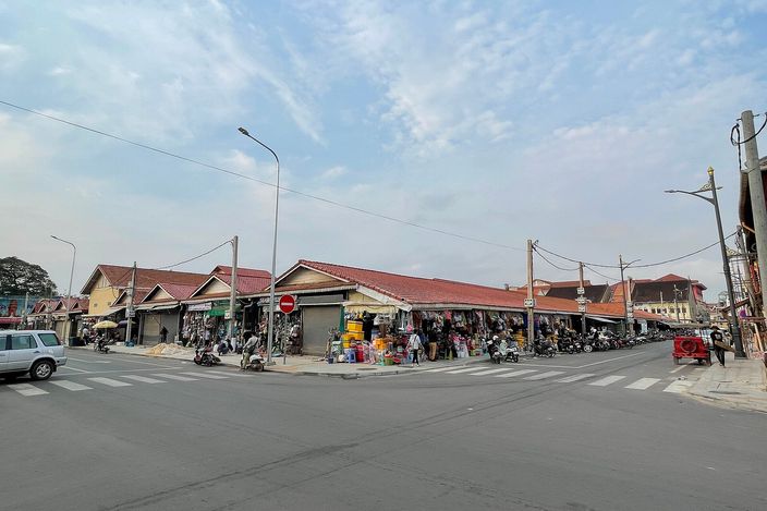The low tile-roofed Old Market (Psar Chas) building of Siem Reap seen across a wide road junction, with motorbikes parked in front and stalls busy under the eaves beneath a pale blue sky.