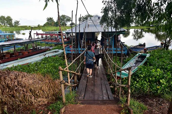A wooden footbridge leads down to a cluster of long, canopied wooden boats moored among water hyacinth on the Tonlé Sap near Kampong Khleang, with tourists walking toward the boats.