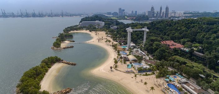 An aerial view of Siloso Beach on Sentosa: a curve of pale sand behind two small engineered headlands and a rock breakwater, palms and low resort buildings behind it, with Singapore's skyline and the gantry cranes of the container port along the horizon.