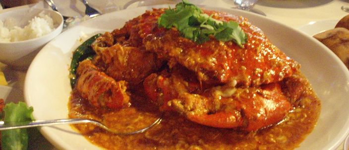 A whole mud crab in its shell coated in a glossy red-orange chilli-crab sauce, plated at a restaurant table with rice and cucumber.