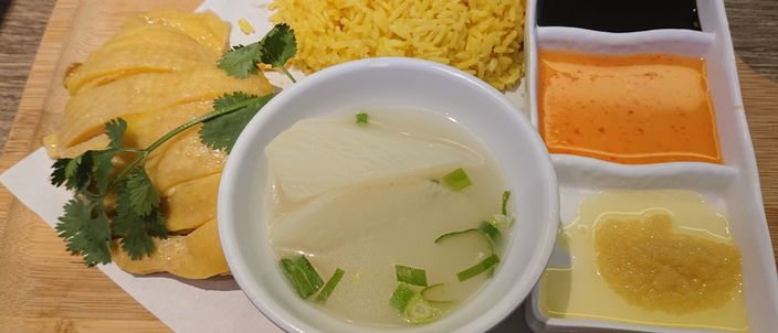 A set of Hainanese chicken rice: pale poached chicken, a mound of chicken-fat rice, a bowl of clear radish soup and a tray of dipping sauces including red chilli.