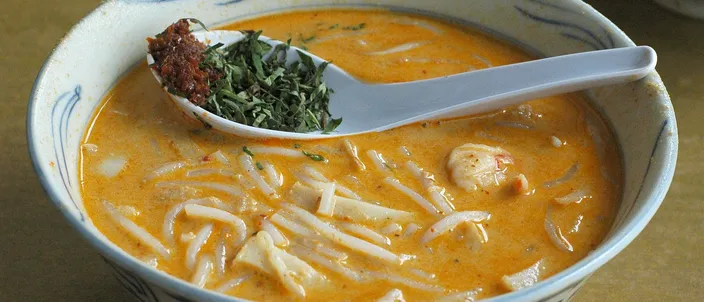 A bowl of Katong laksa: thick rice noodles in an orange coconut-curry gravy, a spoon lifting a topping of torn laksa leaf.