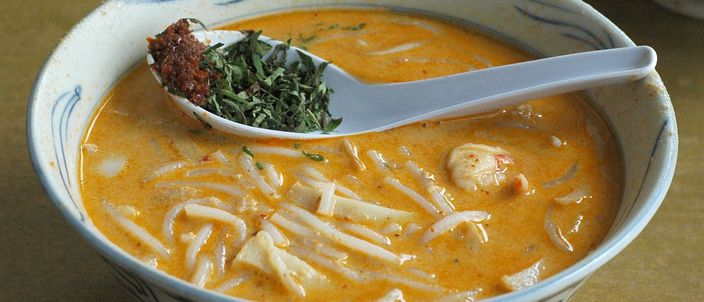 A bowl of Katong laksa: thick rice noodles in an orange coconut-curry gravy, a spoon lifting a topping of torn laksa leaf.