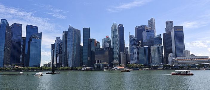 The Singapore Central Business District skyline seen across Marina Bay water, a dense cluster of glass towers under a blue sky with small boats on the water in the foreground