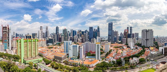 Singapore's financial district towers rising behind red-tiled shophouse roofs and pastel public-housing slabs, seen from above under a bright cloudy sky