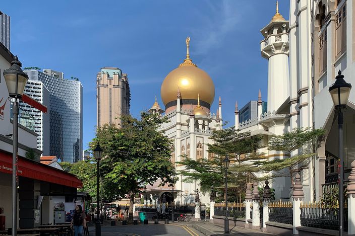 The Sultan Mosque's gilded onion dome and white minaret above a quiet Kampong Glam street, rain trees in front of it and the financial district's towers rising behind