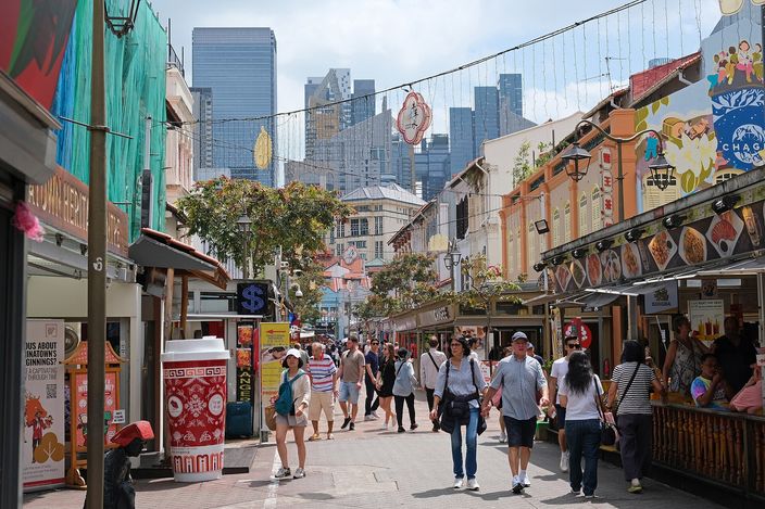 Pedestrians strolling down pedestrianised Pagoda Street in Chinatown, lined with red-and-ochre restored shophouses and hung with decorations, the CBD towers rising behind