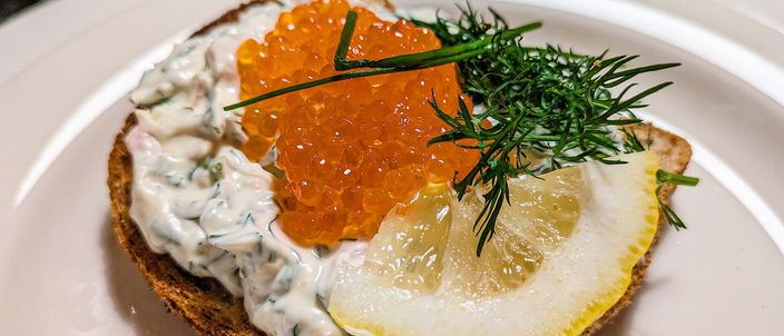 A slice of fried bread topped with a creamy prawn mixture, a mound of orange roe, a sprig of dill and a slice of lemon, on a white plate.