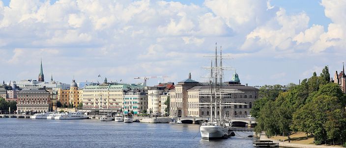 Riddarfjärden waterfront in summer: the white sailing-ship af Chapman moored off Skeppsholmen, ochre facades of the Grand Hôtel and Nationalmuseum behind, Gamla Stan spires to the left under a bright cloud-dappled sky.