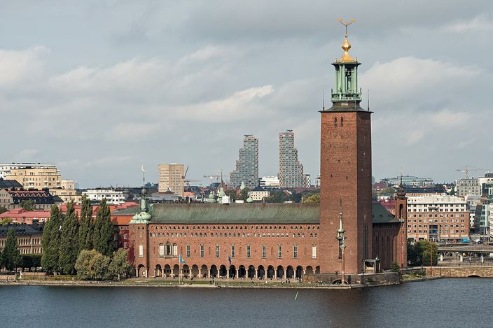 A dark red brick building with an arcaded waterfront terrace and a tall square tower topped by a green lantern and a gilded finial, seen across a wide stretch of water, with trees to the left and a modern high-rise skyline in the distance under a cloudy sky.