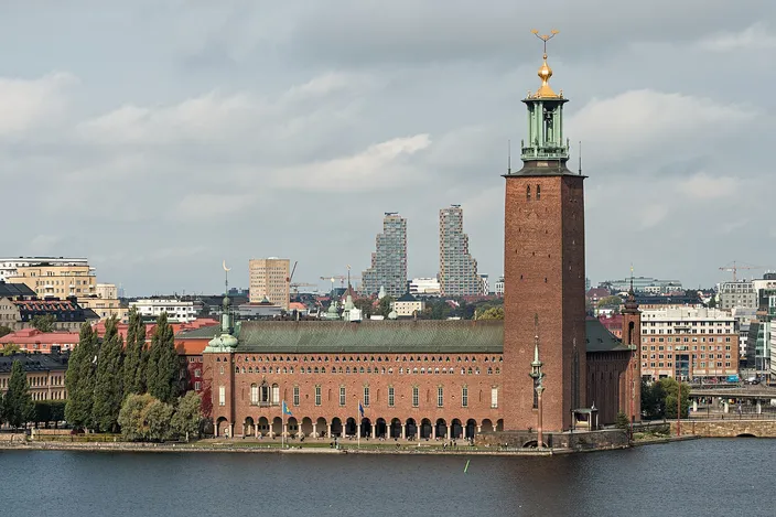 A dark red brick building with an arcaded waterfront terrace and a tall square tower topped by a green lantern and a gilded finial, seen across a wide stretch of water, with trees to the left and a modern high-rise skyline in the distance under a cloudy sky.